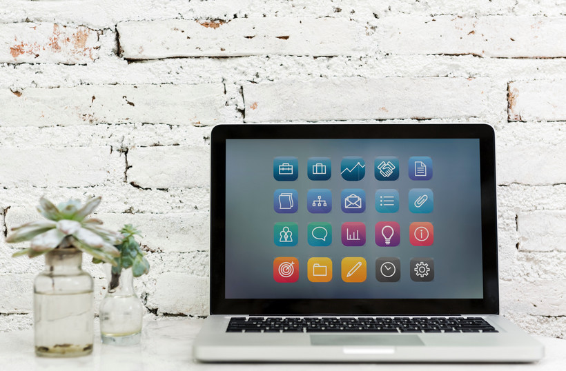 Auf dem Bild ist ein aufgeklappter Laptop vor der weißen Backsteinwand zu sehen. Der Bildschirm zeigt verschiedene bunte Icons mit Werkzeugsymbolen. Links vom Laptop stehen zwei Vasen mit grünen Pflanzen.