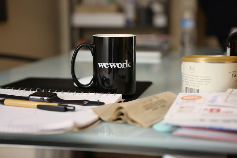 Das Foto zeigt einen Schreibtisch, auf dem ein Schreibblock mit einem Kugelschreiber und einer Armbanduhr abgebildet sind. Der Fokus liegt auf der dahinter stehenden schwarzen Tasse mit dem Druck "wework".