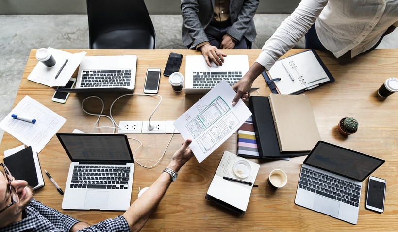 Ein Schreibtisch mit Laptops, Kaffee-Bechern und drei Menschen, die gerade zusammenarbeiten Von oben fotografiert: Ein Schreibtisch, an dem drei Personen an den Laptops arbeiten. Eine Person reicht der anderen gerade ein Dokument über.