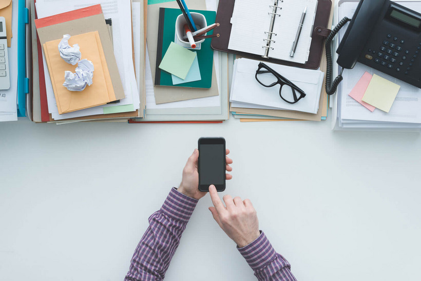 Man sieht eine Hand, die ein Smartphone hält. Im Hintergrund sieht man einen Schreibtisch mit Utensilien.