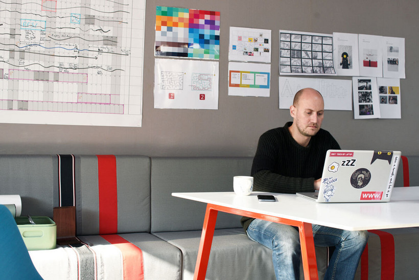 Digital erfolgreich: 5 Methoden, die Unternehmen von Start-ups lernen können - Ein Mann sitzt in einem modernen Büro vor einem Tisch Auf dme Bild sieht man einen Mann mit schwarzen Pullover und Glatze, er arbeitet an einem Schreibtisch in einem modernen und jungen Büro. Hinter ihm sind an der Wand Schaugrafiken und Poster.