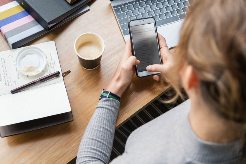 Die Arbeit eines Digital Change Manager Das Bild zeigt eine Frau an einem Handy. Sie sitzt an einem Schreibtisch mit Block und Laptop und arbeitet.